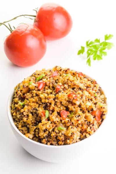 A bowl of cooked quinoa with herbs and spices sits in front of tomatoes on the vine and fresh green parsley.