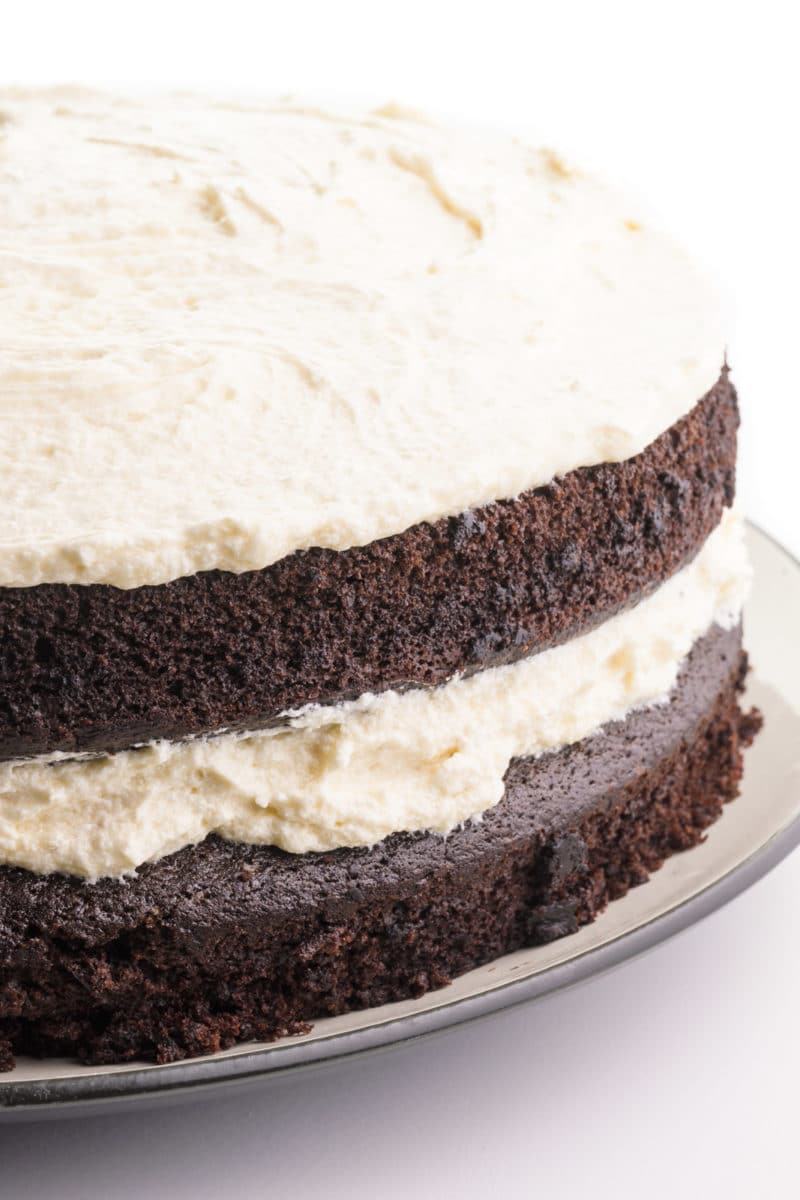 A vegan Suzy Q Cake sits on a plate with frosting on top and between the layers, showing the chocolate cake along the edges.