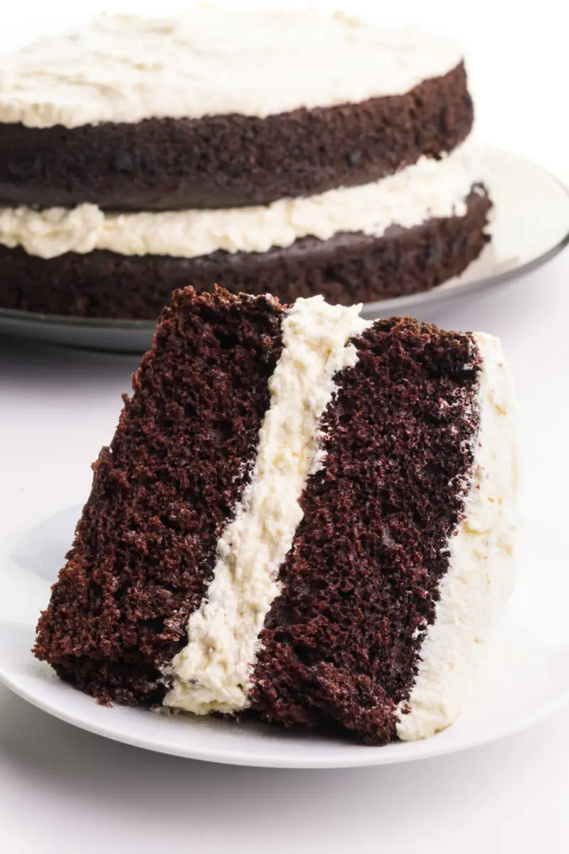 A slice of chocolate cake sits on its side, showing lots of fluffy frosting. The rest of the cake sits on a plate behind it.