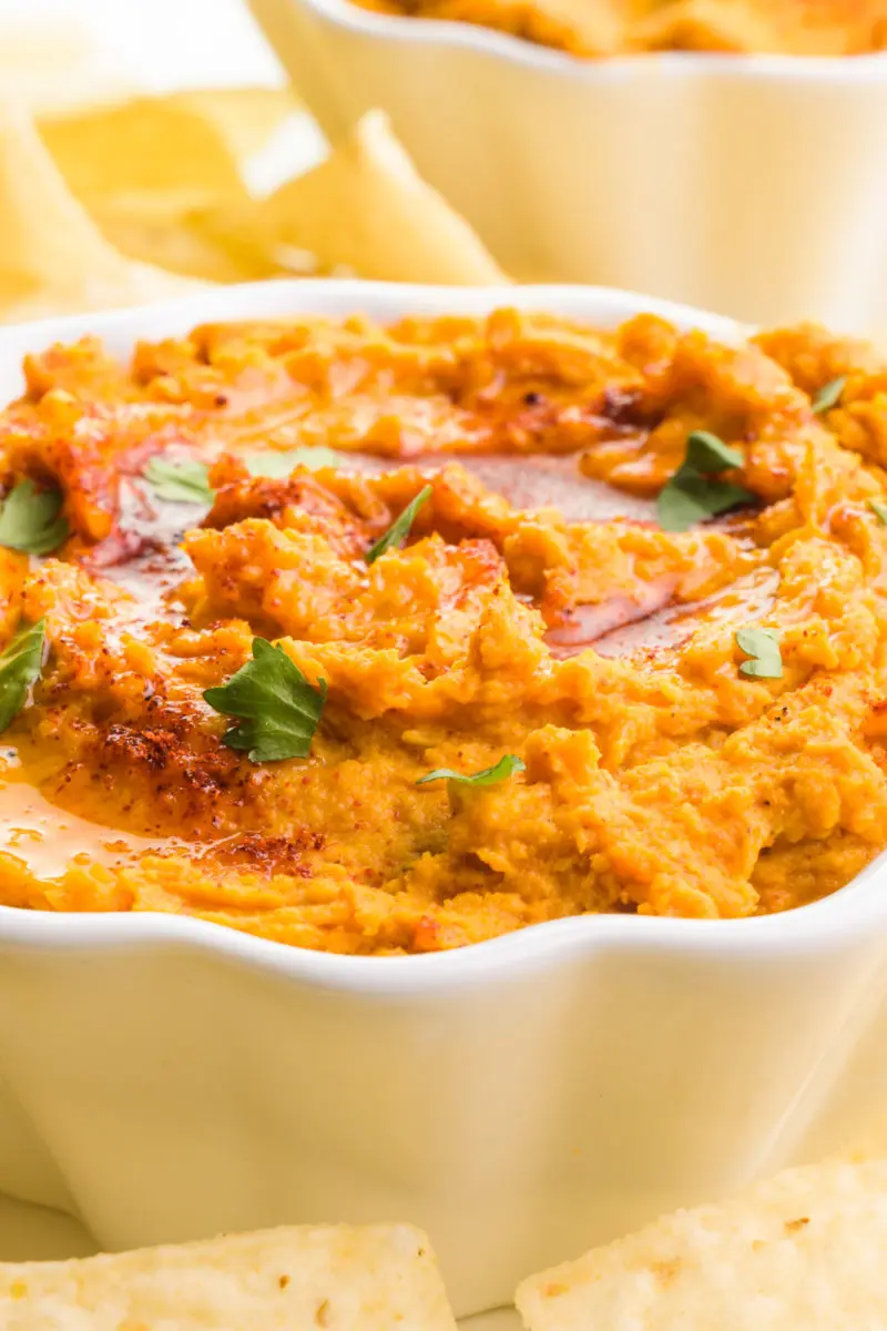 A close-up shot of sweet potato hummus in a bowl. It's topped with chopped cilantro, olive oil, and paprika. There's another bowl and tortilla chips in the background.
