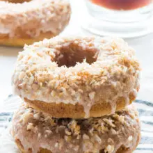 A stack of toasted coconut donuts sits on a kitchen towel with another donut in the background next to a cup of tea