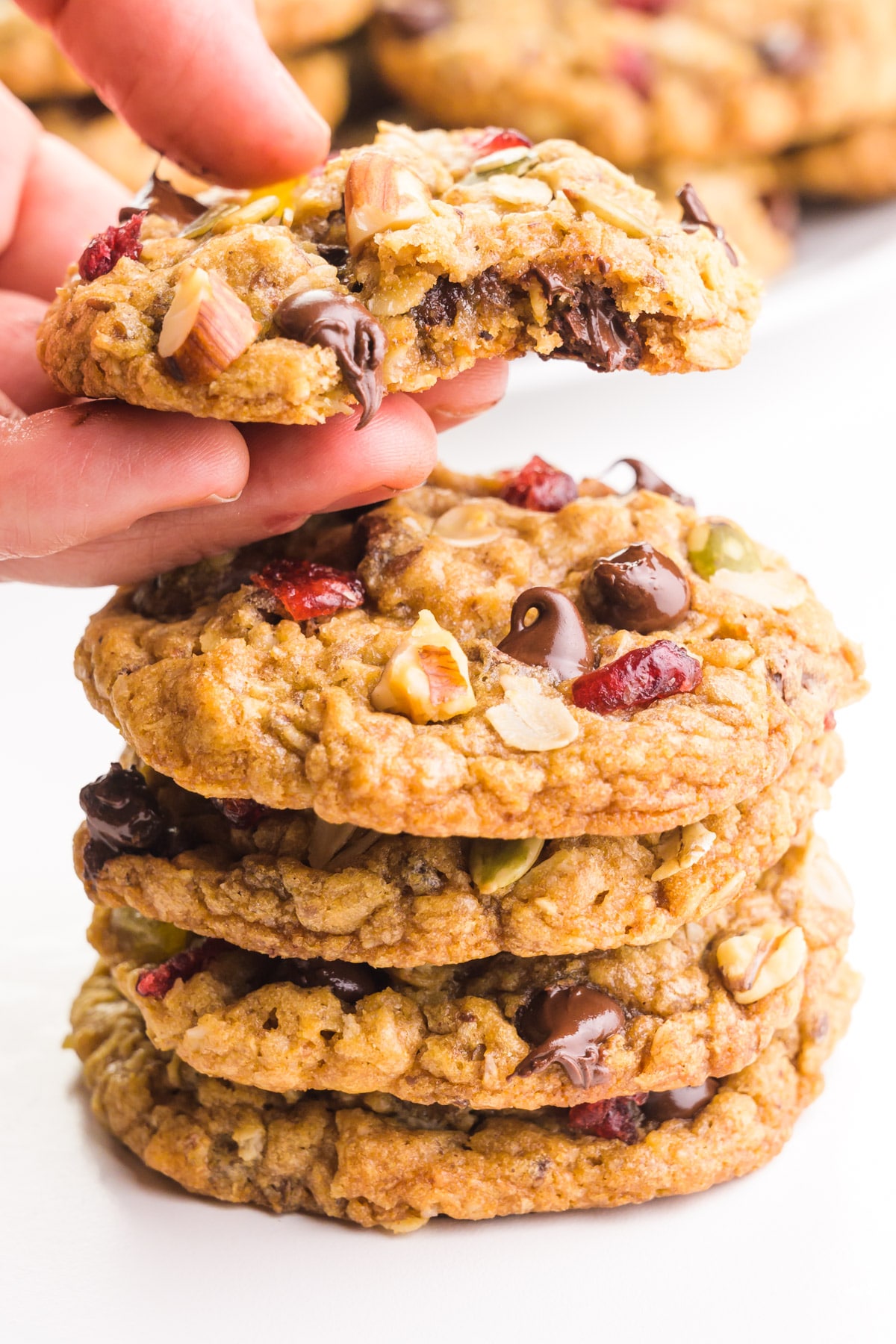 A hand holds a trail mix cookie with a bite taken out, showing lots of melty chocolate chips. It's hovering over a stack of cookies and there are more cookies in the background.