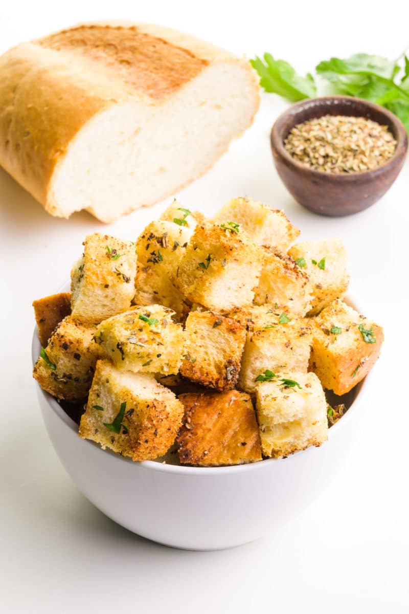 A bowl of croutons sits in front of a bowl of seasonings, herbs, and bread.