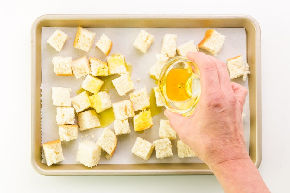 A hand holds a small bowl of olive oil, drizzling over bread crumbs in a pan.