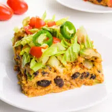 A slice of enchilada casserole sits on a plate. There are cherry tomatoes in the background.