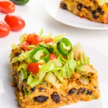 A slice of vegan enchilada casserole sits on a plate. There's another slice on a plate behind it along with some veggies beside it.