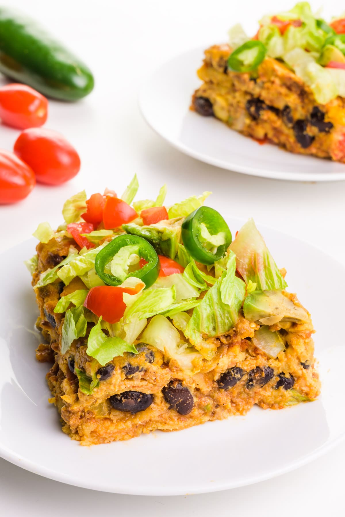 A slice of vegan enchilada casserole sits on a plate. There's another slice on a plate behind it along with some veggies beside it.