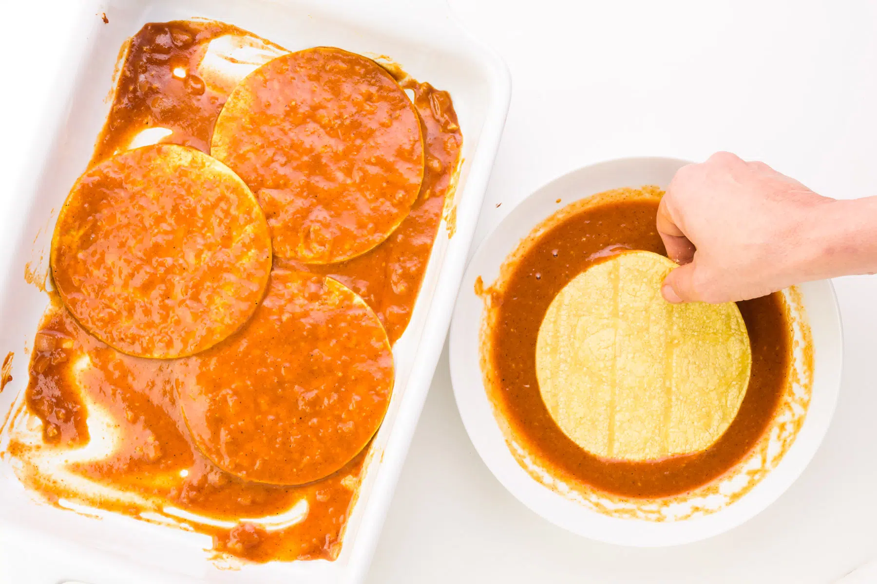 A hand is dipping tortillas in enchilada sauce and arranging them in a casserole dish.