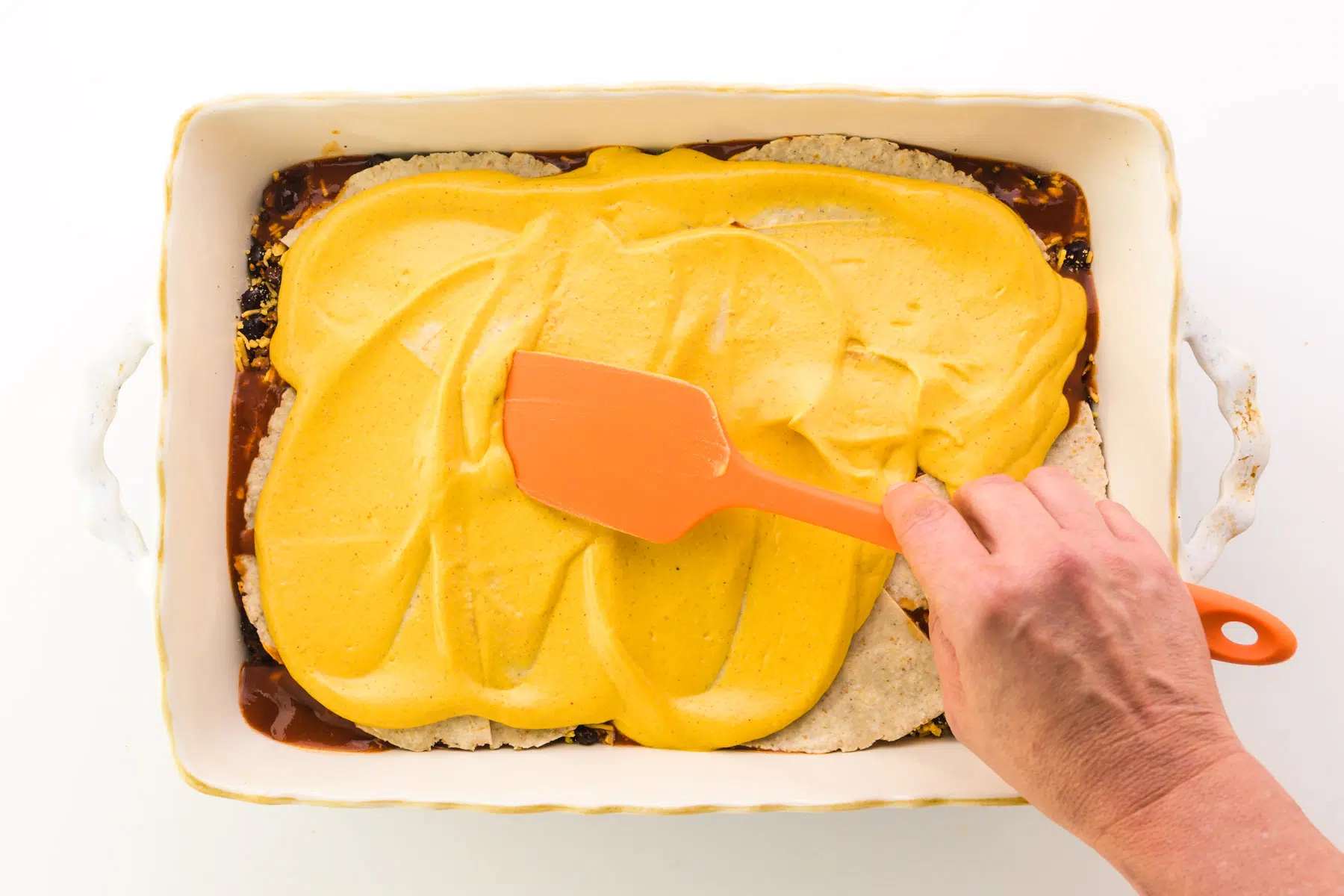 A hand holds an orange spatula, using it to spread cheese sauce over a casserole.