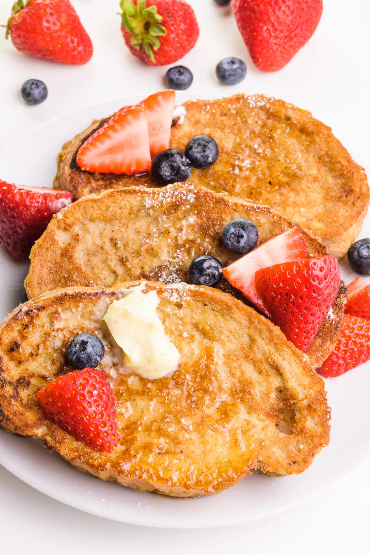 Three slices of French toast have fruit on top, some butter, and powdered sugar. There is fresh fruit situated around the plate.