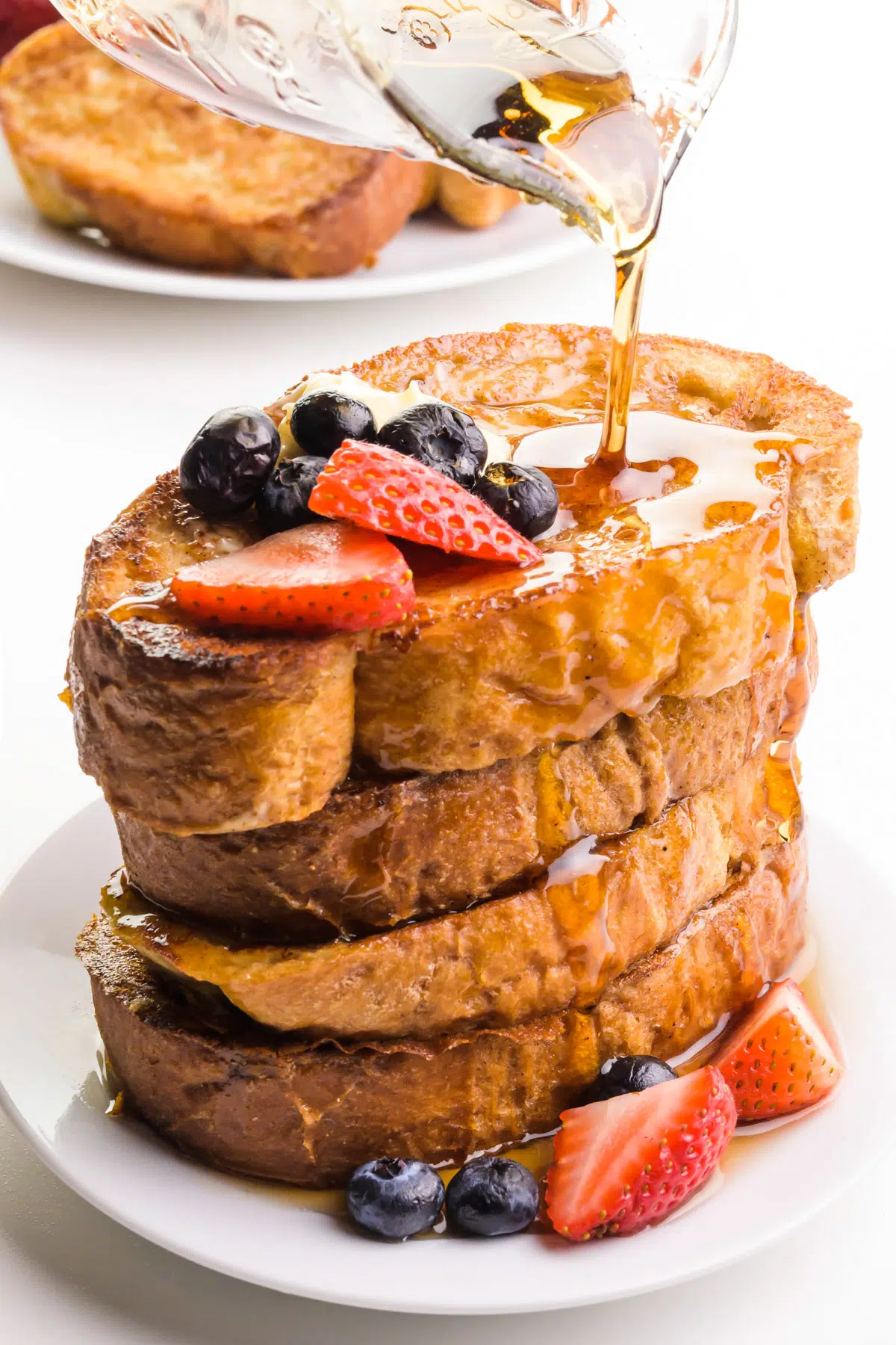 Syrup is being poured over a stack of vegan French toast. There's a plate with more French toast in the background.