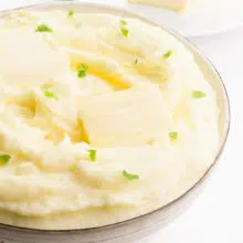 Looking down on a bowl full of vegan mashed potatoes. There is melted butter on top with parsley flakes and a plate with more vegan butter in the background.