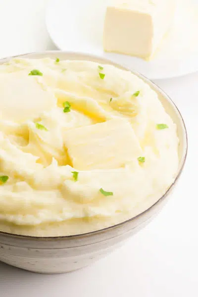Looking down on a bowl full of vegan mashed potatoes. There is melted butter on top with parsley flakes and a plate with more vegan butter in the background.