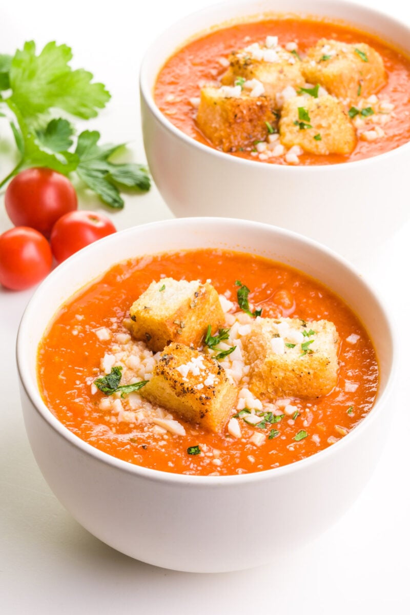 Two bowls hold tomato soup with croutons next to fresh tomatoes.