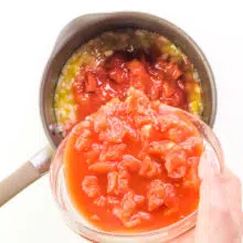 A hand holds a bowl of tomatoes, pouring it into a saucepan with cooked onions.