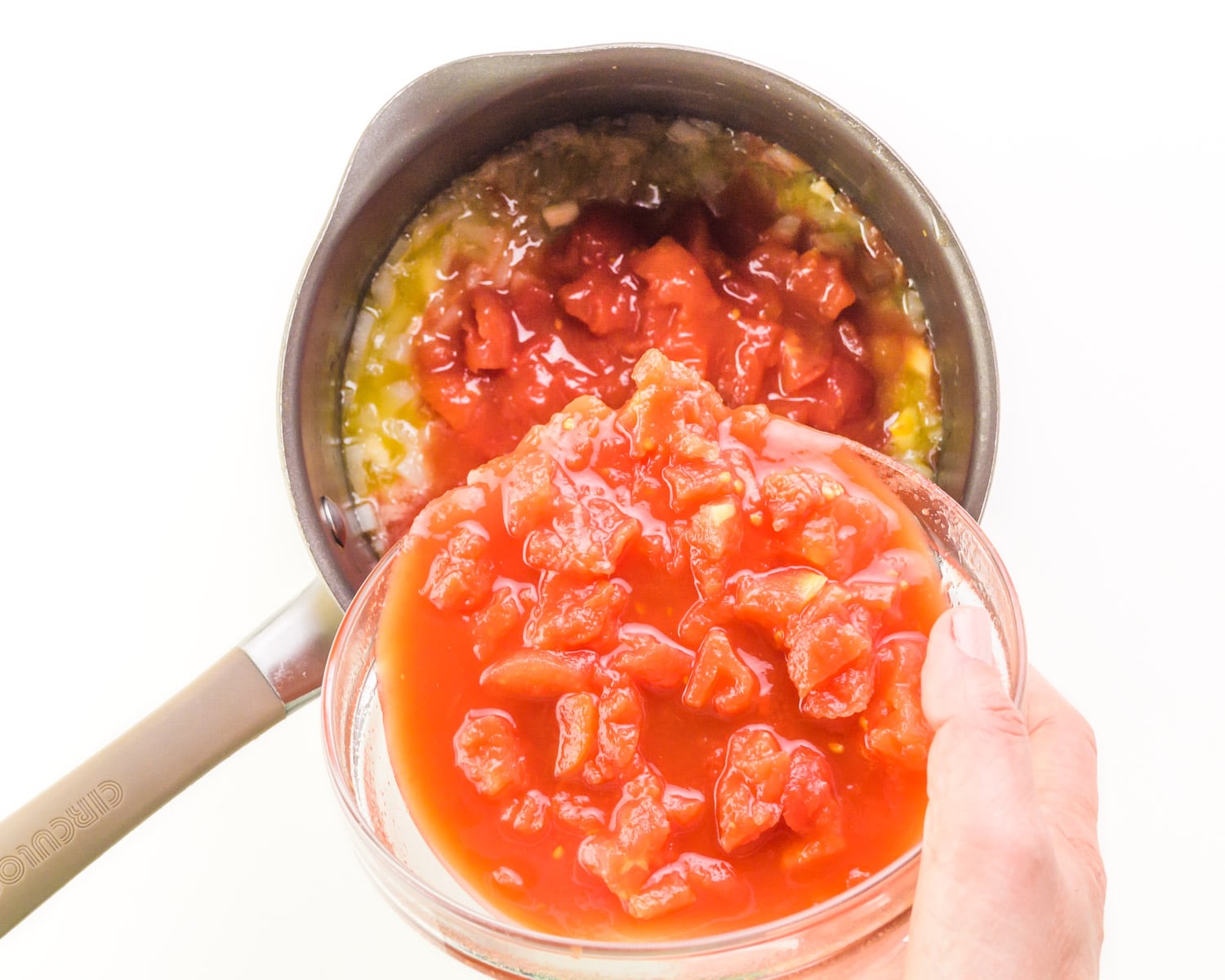 A hand holds a bowl of tomatoes, pouring it into a saucepan with cooked onions.