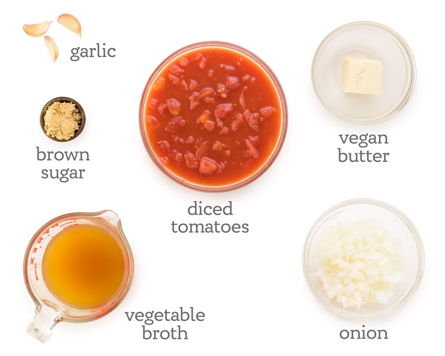 Ingredients are laid out on a table. The labels next to them read, chopped onions, vegan butter, diced tomatoes, garlic, brown sugar, and vegetable broth.
