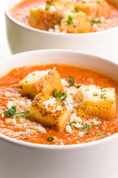 A bowl of vegan tomato soup has croutons and herbs on top. There is another bowl in the background.