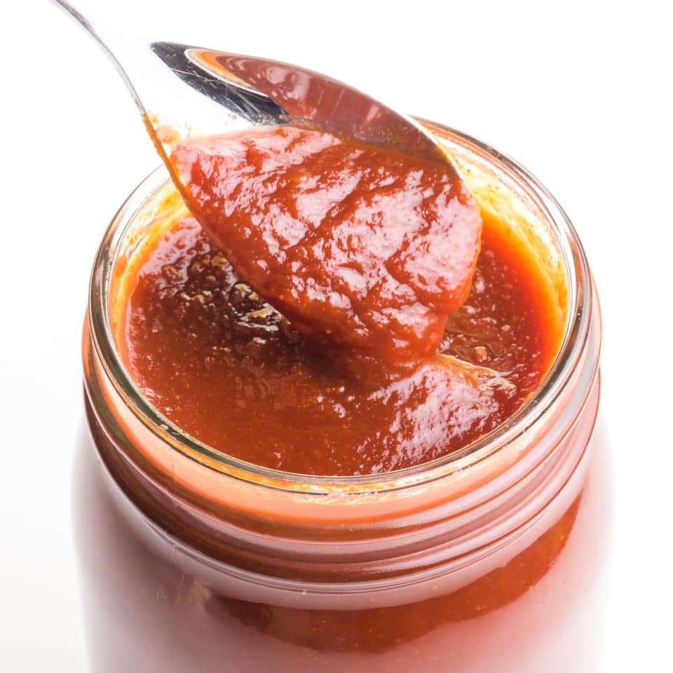 A closeup of a spoon with bbq sauce hovering over a jar full of the sauce.