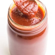A spoon holds vegan bbq sauce over a glass jar full of the sauce.