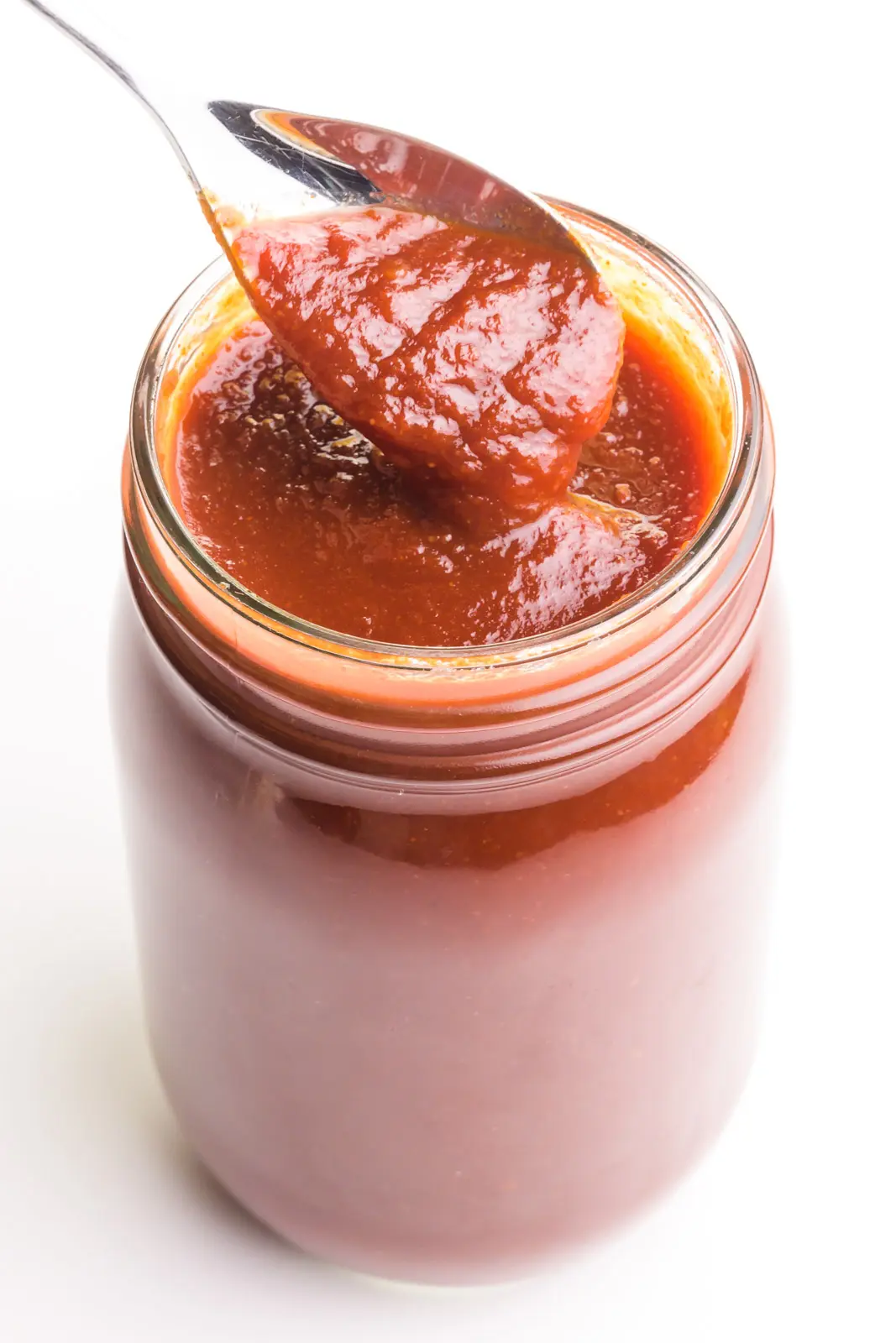 A spoon holds vegan bbq sauce over a glass jar full of the sauce.