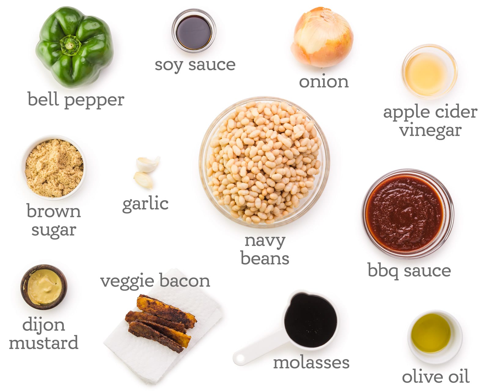 Ingredients are laid out on a table. The labels next to them read, onion, apple cider vinegar, bbq sauce, olive oil, molasses, navy beans, veggie bacon, dijon mustard, brown sugar, bell pepper, garlic, and soy sauce.