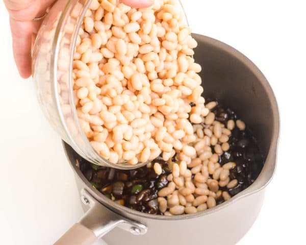 A hand holds a bowl, pouring navy beans into a pan with BBQ sauce mixture.