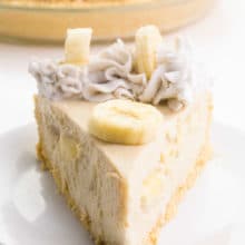 A slice of vegan banana cream pie sits on a plate. It has banana slices and coconut whipped cream on top. The rest of the pie is behind it.