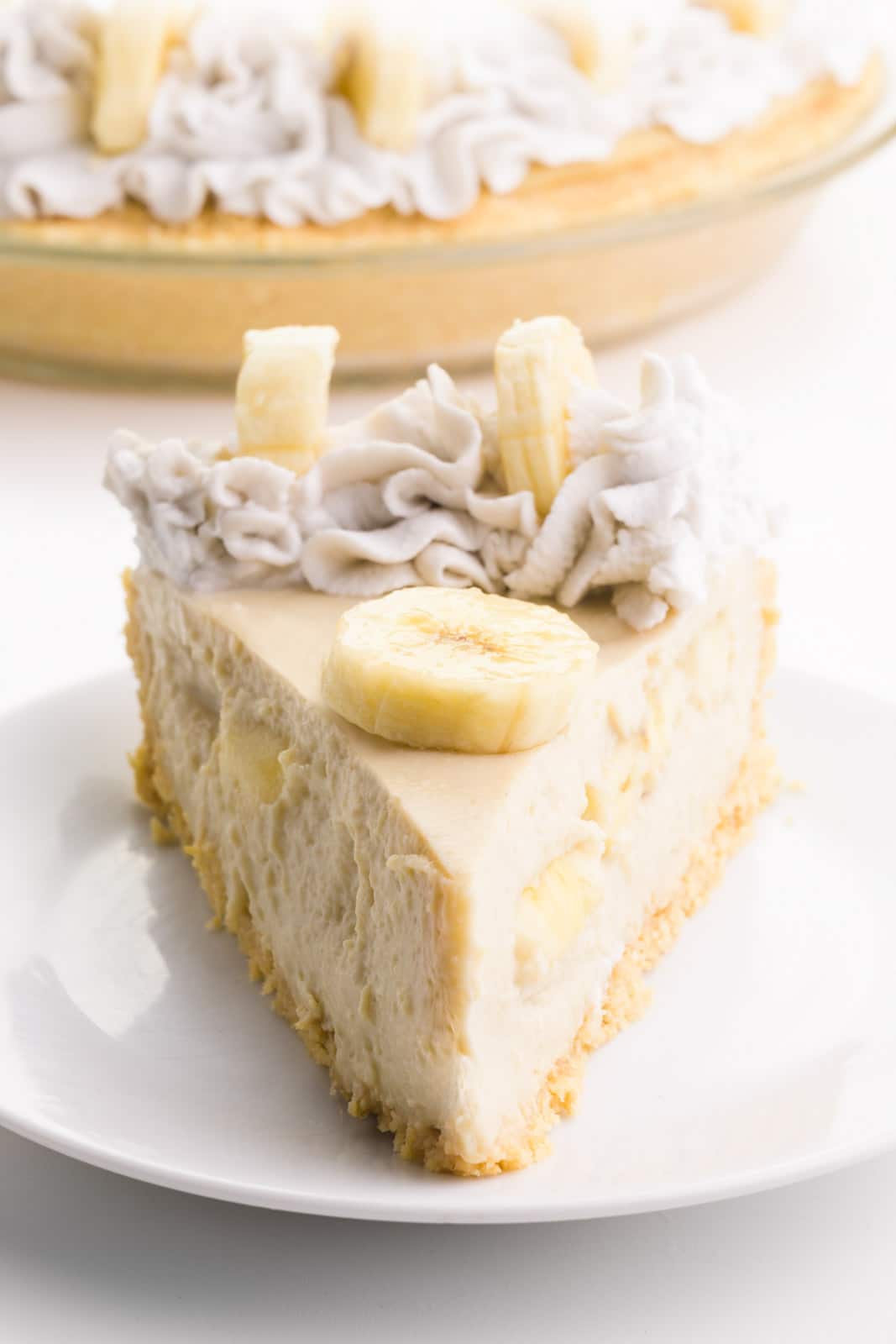 A slice of vegan banana cream pie sits on a plate. It has banana slices and coconut whipped cream on top. The rest of the pie is behind it.