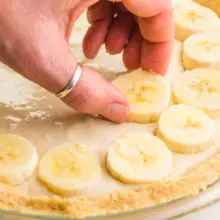 A hand is placing sliced bananas on a pie filled with vanilla pudding.