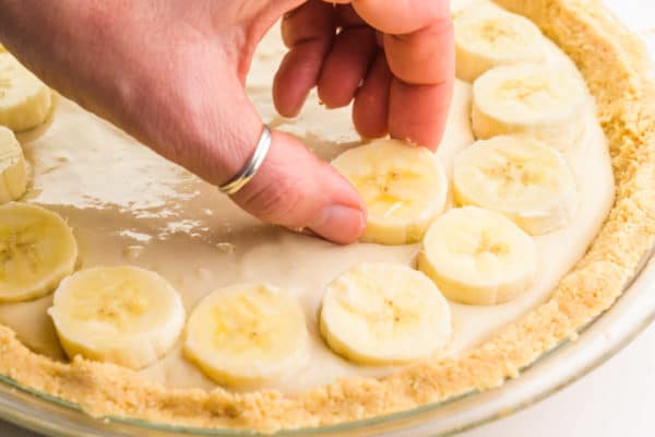 A hand is placing sliced bananas on a pie filled with vanilla pudding.