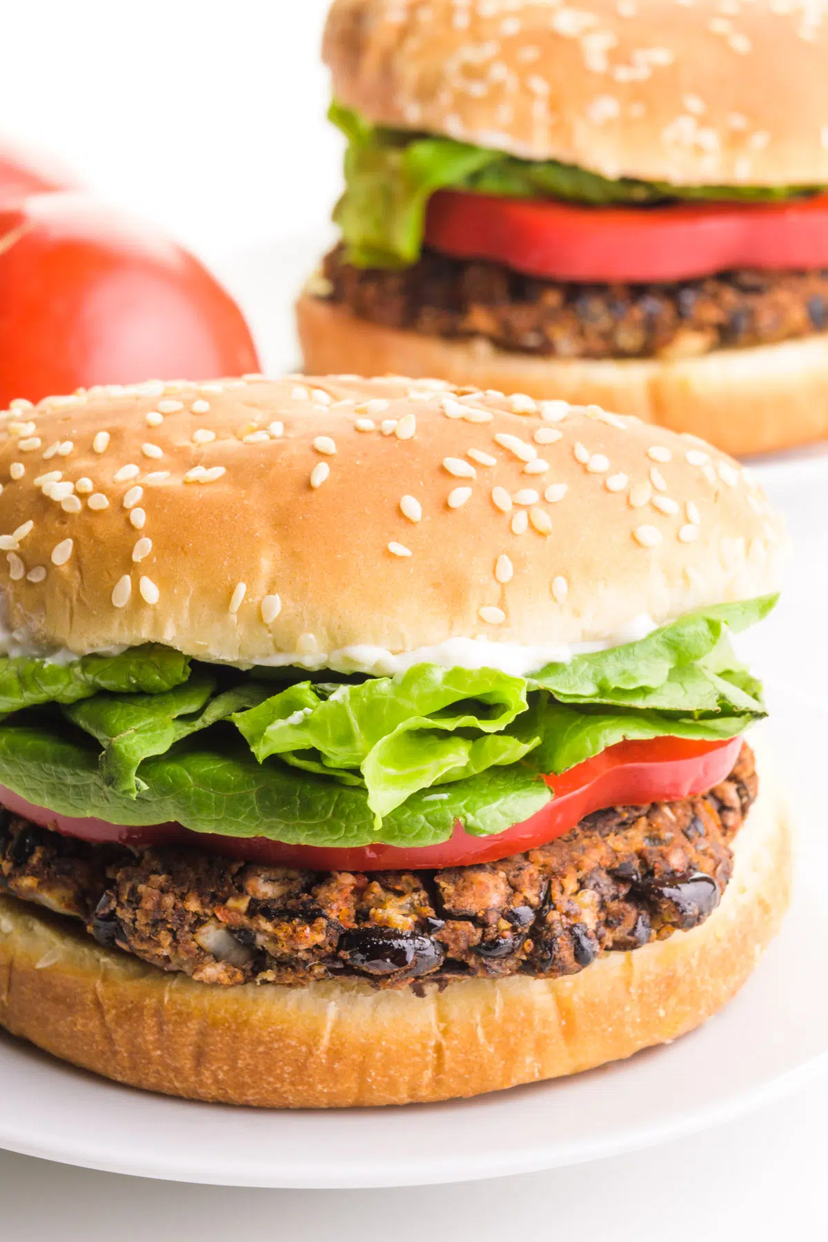 A vegan black bean burger sits on a bun with toppings like lettuce. There's another burger in the background and a tomato.
