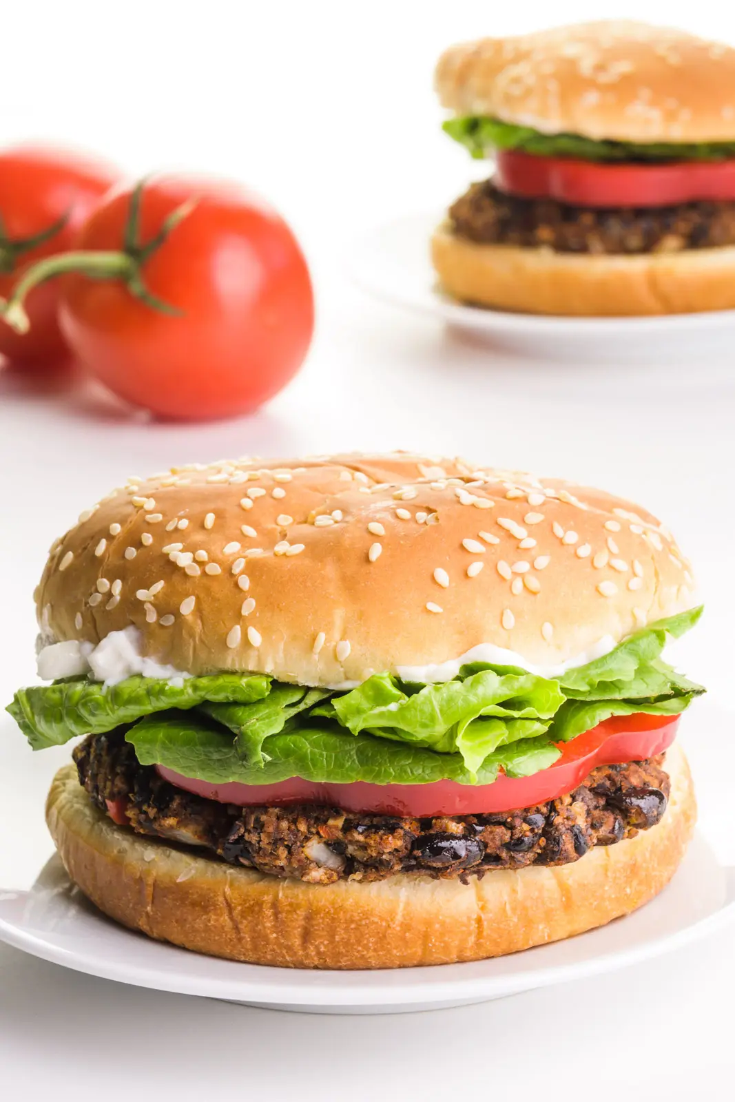 A vegan black bean burger sits on a plate. There's another burger and two tomatoes in the background.