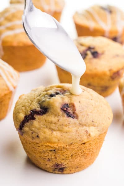A spoon hovers over a muffin, preparing to drizzle it with glaze. There are more muffins in the background.