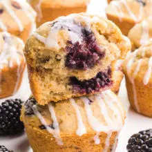 A closeup of a blackberry muffin with a bite taken out. It is sitting on another muffin. There are blackberries and more muffins around it.