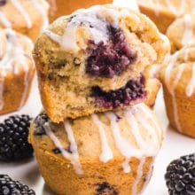 A blackberry muffin with a bite out of it sits on top of another muffin. There are fresh blackberries and more muffins around the stack.