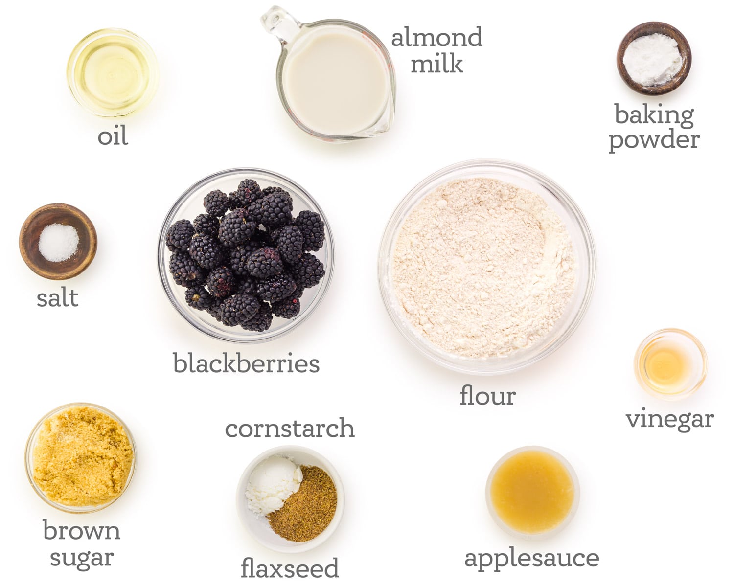 Ingredients are displayed on a white counter. The labels next to them read, baking powder, vinegar, flour, applesauce, flaxseed, cornstarch, brown sugar, blackberries, salt, oil, and almond milk.
