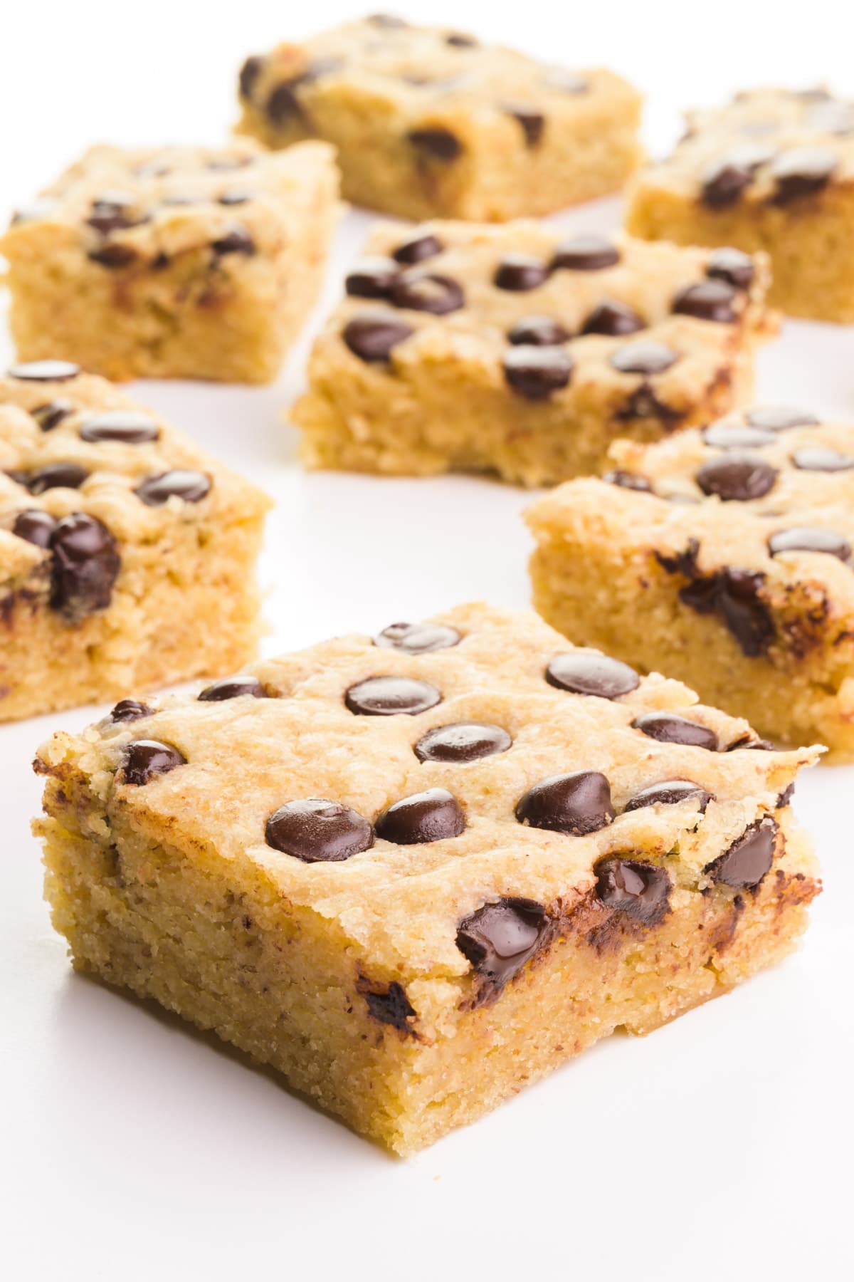 Several slices of vegan blondies have chocolate chips on top. They are displayed on a white table top.