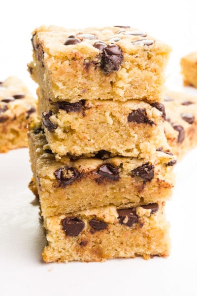 A stack of vegan blondies shows chocolate chips on top and on the sides. There are more slices in the background.