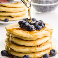 Syrup is being poured over a stack of pancakes with fresh blueberries on top and on the sides. There's another stack of pancakes and more blueberries in the background.