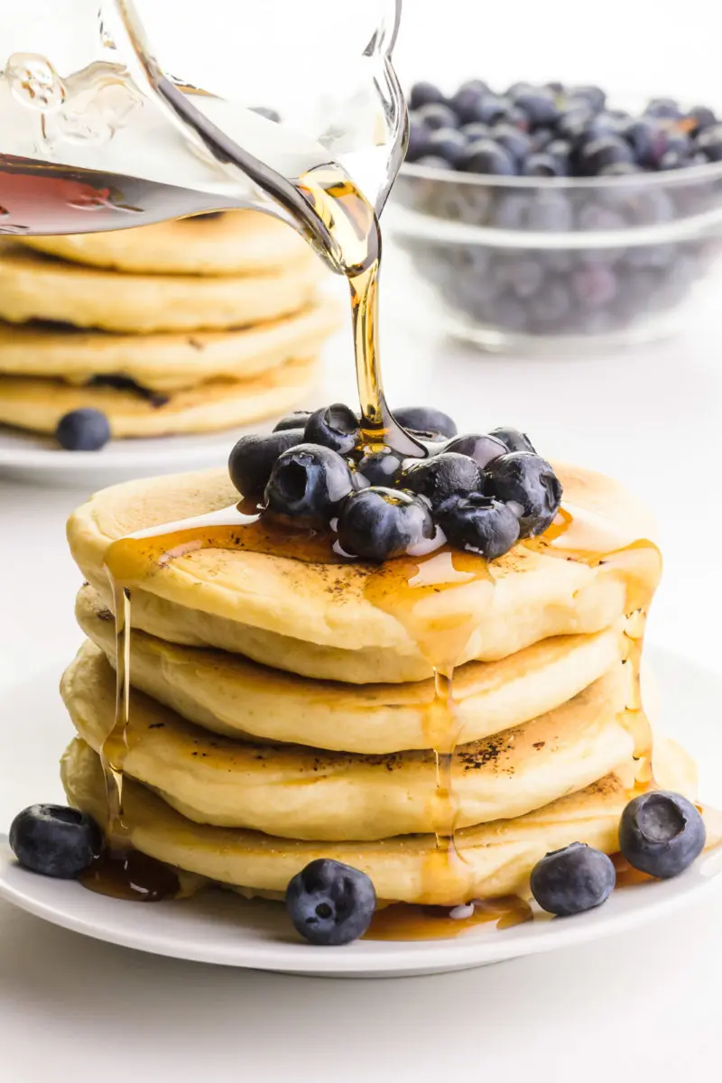 Syrup is being poured over a stack of pancakes with fresh blueberries on top and on the sides. There's another stack of pancakes and more blueberries in the background.