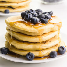 A stack of vegan blueberry pancakes has fresh blueberries and maple syrup on top and on the sides.