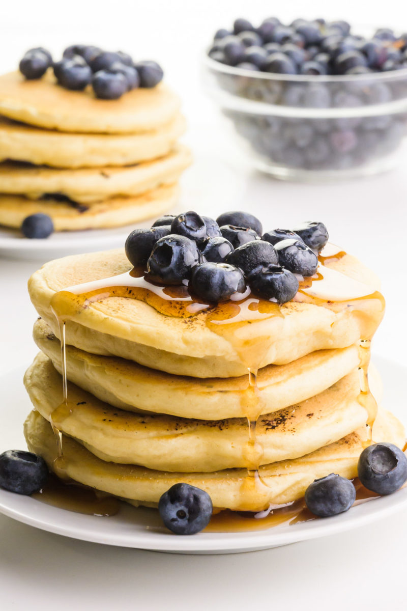 A stack of vegan blueberry pancakes sits on a plate. There are blueberries on top and beside the pancakes and drizzles of syrup coming down the sides. There are more pancakes and blueberries in the background.
