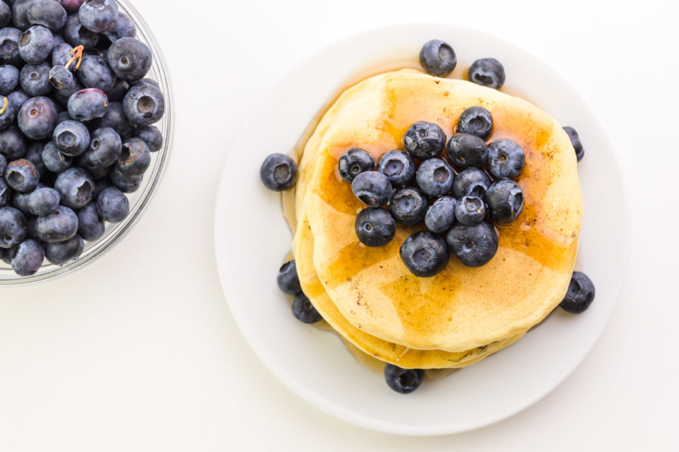 Looking down on a stack of pancakes with blueberries on top and beside it sitting beside a bowl of fresh blueberries.