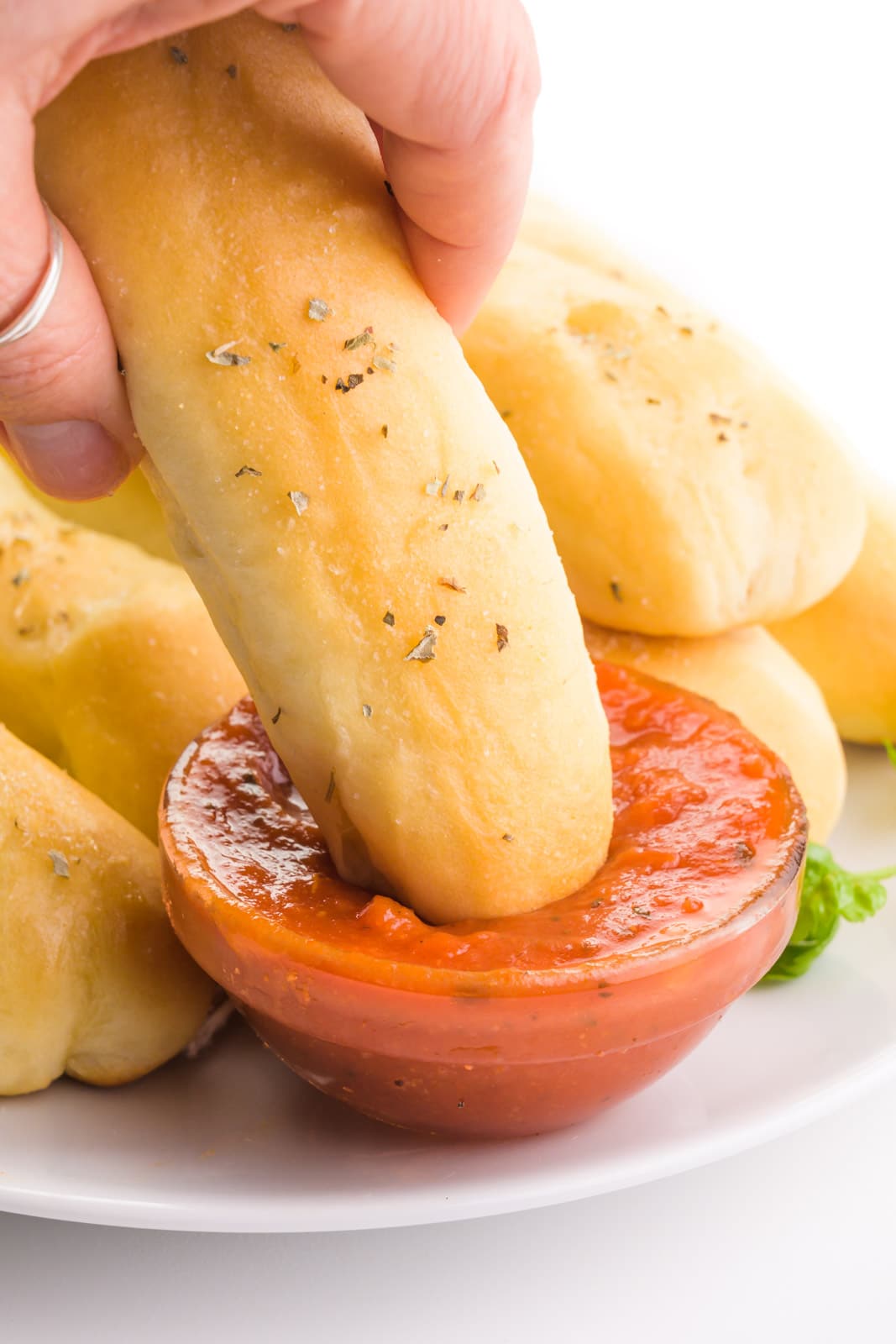 A hand holds a breadstick, dipping it in marinara sauce. There are more breadsticks in the background.