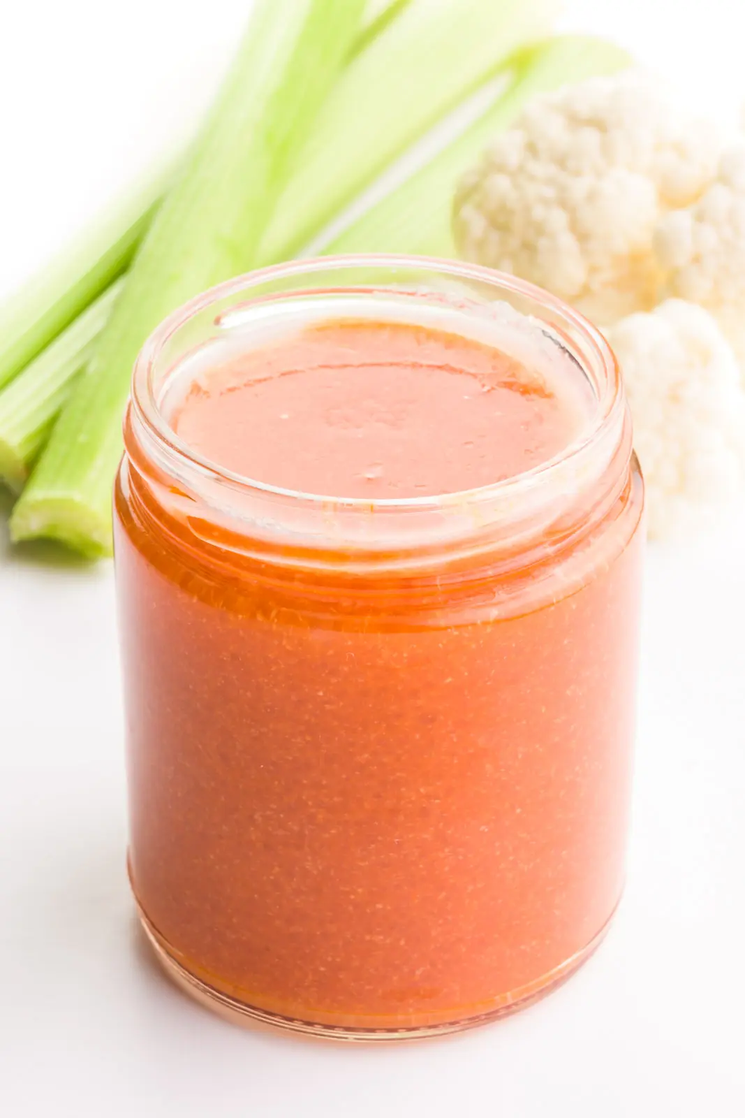 Vegan buffalo sauce is in a glass jar. There are stalks of celery and cauliflower in the background.