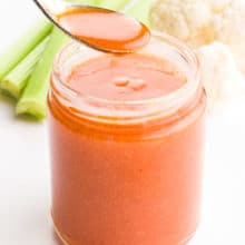 A spoon full of vegan buffalo sauce hovers over the rest of the jar. There are celery sticks and cauliflower florets in the background.
