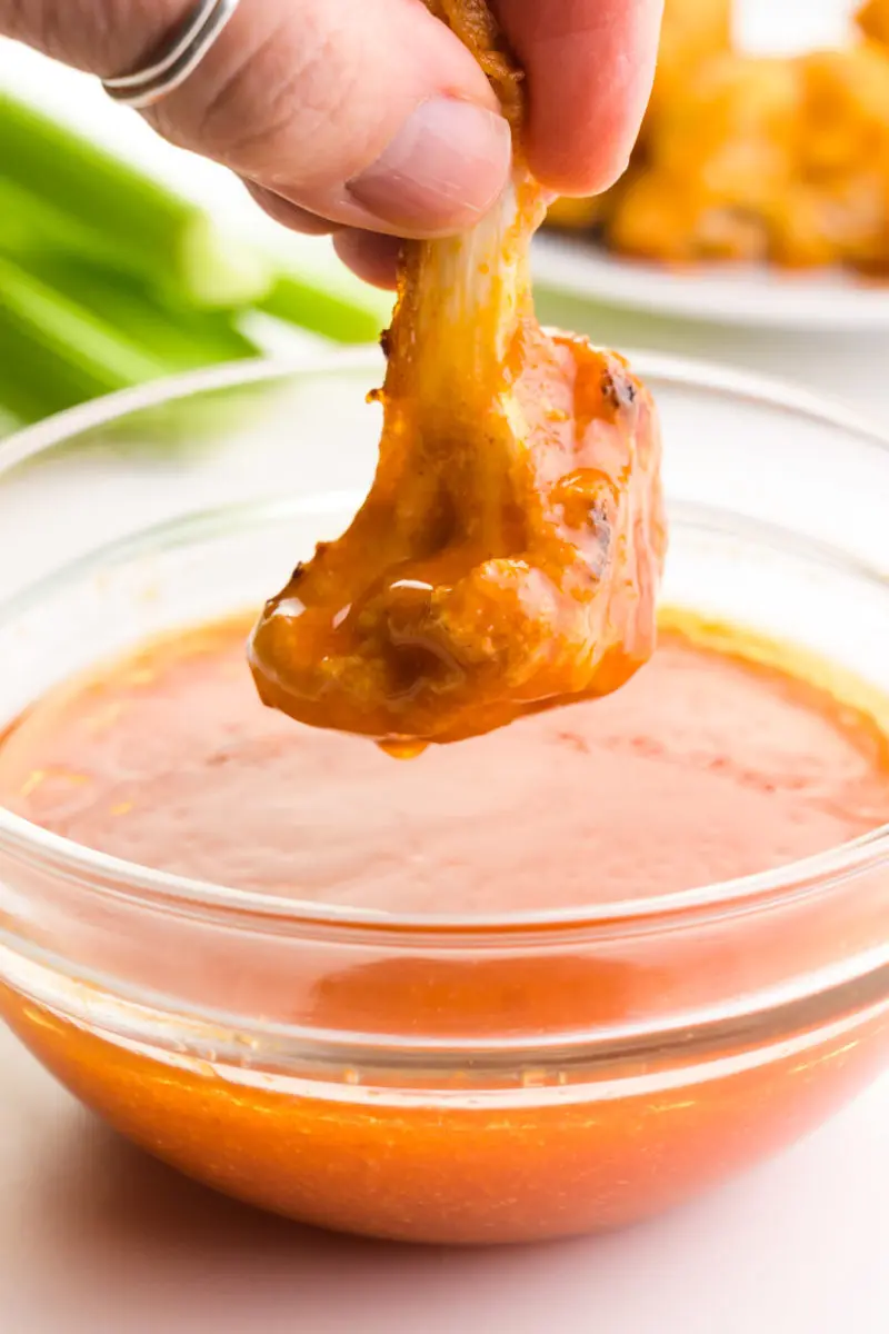 A hand holds a cauliflower wing after dipping it in vegan buffalo sauce. There are celery sticks and more cauliflower wings in the background.