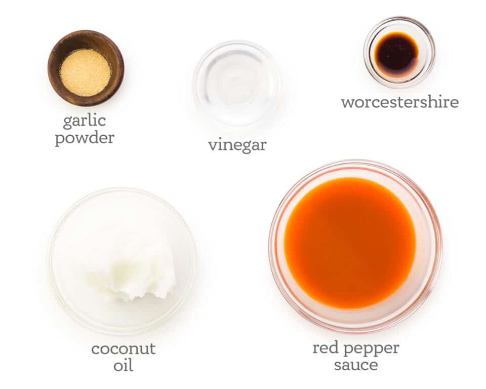 Several ingredients sit on a white counter. The labels next to them read, Garlic powder, vinegar, Worcestershire, Red Pepper Sauce, and Coconut Oil.