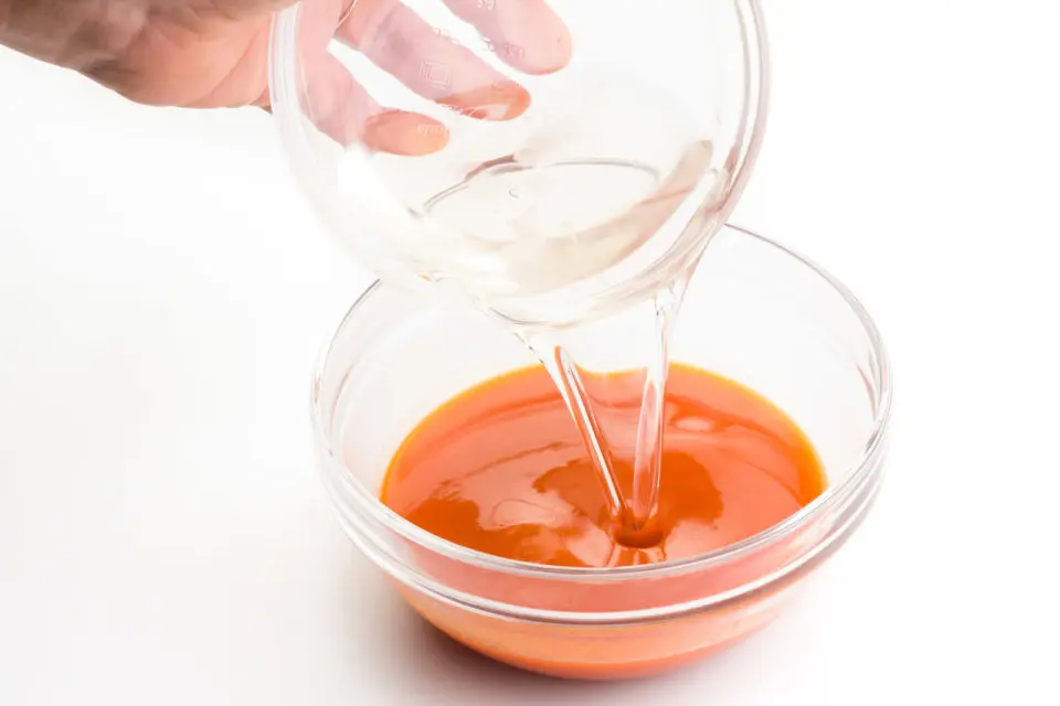 A. hand holds a bowl of melted coconut oil, pouring it into another bowl with red pepper sauce.